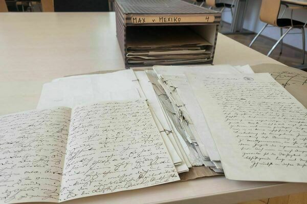 Picture of a box in an archive with the label "Max von Mexiko." There are a number of documents open on the table.