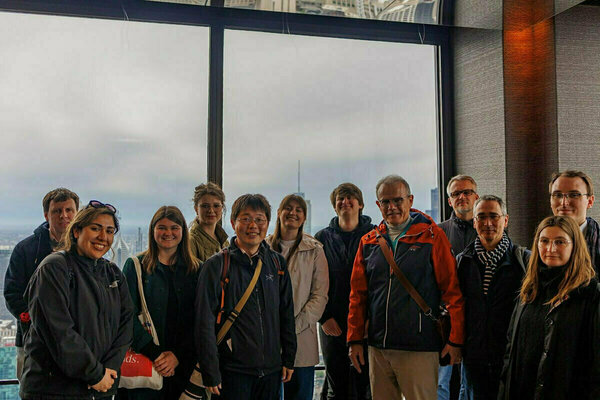 A group of eleven people stands before large windows offering a cloudy cityscape view.
