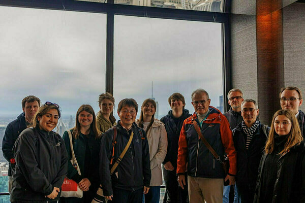 A group of eleven people stands before large windows offering a cloudy cityscape view.