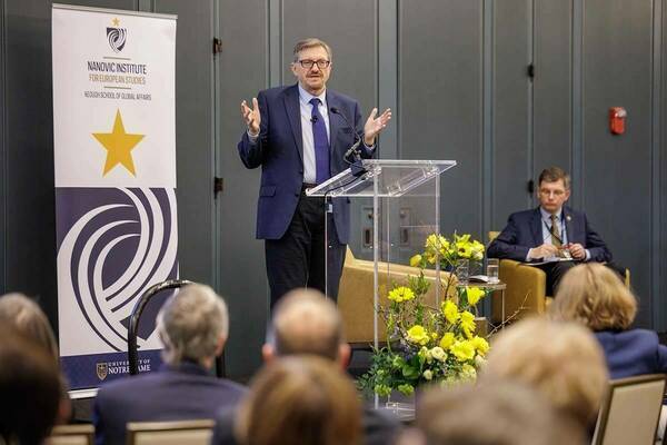 A man in a navy suit and blue tie gestures while speaking at a clear podium in front of an audience.  A banner for the Nanovic Institute for European Studies at the University of Notre Dame stands to the left.  Yellow flowers are arranged near the podium. Another man in a suit sits to the right.